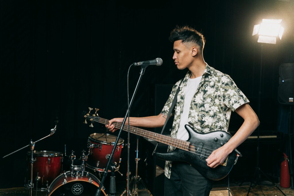 A male musician in a floral shirt playing a bass guitar on stage during a live performance.